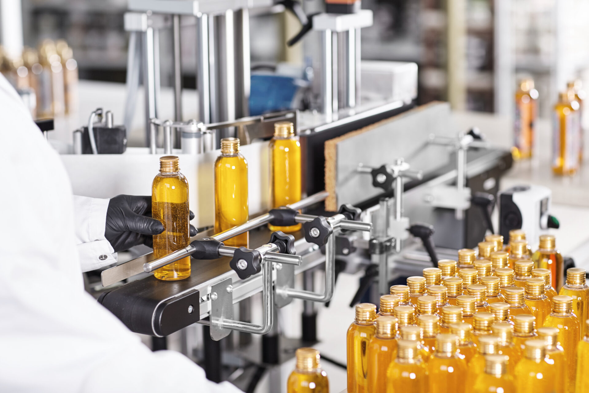 Rear view of unrecognizable factory worker in gown and gloves controlling production process of manufacturing cosmetics at modern laboratory, taking bottle of golden yellow liquid from conveyor line